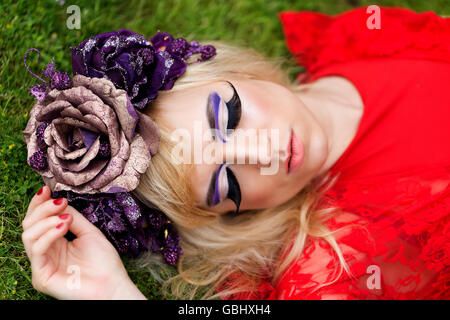 Blonde Modells, feinen Gesichtszügen, purpurrote Blume Krone, geflügelte Eyeliner, riesige Wimpern, rotes Spitzenkleid, schlafen auf dem Rasen liegen Stockfoto