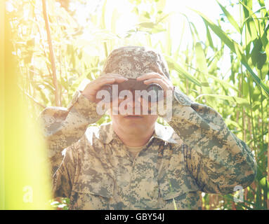 Ein Soldat Mann sucht durch ein Fernglas in den Wäldern außerhalb mit einer einheitlichen Schutz, Krieg oder Jäger Konzept auf. Stockfoto