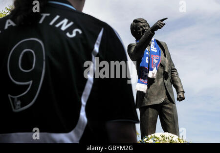 Ein Mann, der ein Newcastle United-Hemd trägt, sieht an, als neben einer Statue von Bobby Robson vor dem Fußballverein von Ipswich Town Blumen und Schals zu sehen sind, in Erinnerung an den Fußballmanager, der heute verstorben ist, Ipswich. Stockfoto