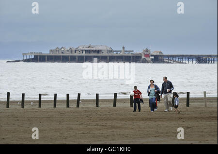 An einem grauen und bewölkten Tag wandern die Menschen entlang des Weston-Super-Mare Strandes Somerset mit dem Birnbeck Pier im Hintergrund. Stockfoto