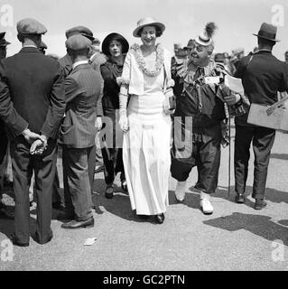 Eine gut gekleidete Dame bei den Rennen in Royal Ascot zeigt ihre Belustigung, als sie von einem Clown in vollem Kostüm begleitet wird, während sie sich durch die Menge macht. Stockfoto