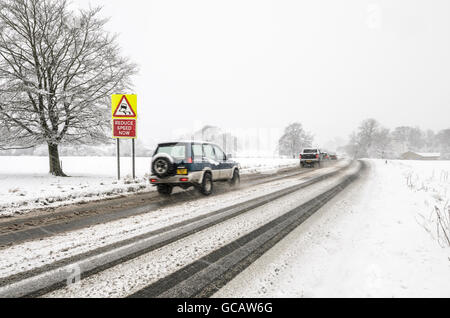 Autos fahren vorbei an eine glatte Oberfläche, die Warnung Straßenschild, während es mit Schnee auf dem Boden schneit. Stockfoto