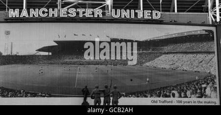 Ein Plakat an der Außenseite des Old Trafford Stadions von Manchester United, das 100 Jahre Old trafford feiert, ebenfalls abgebildet ist die Statue von Denis Law, Sir Bobby Charlton und George Best und die Statue von Sir Matt Busby (im Hintergrund in der Mitte) Stockfoto