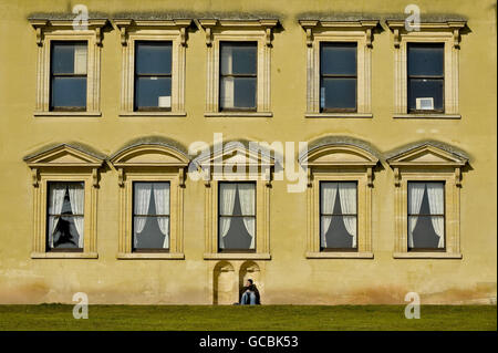 Ein Mann sitzt in einer Aussparung unter den Fenstern auf der Südseite des Ashton Court Mansion, Bristol, aus dem 17. Jahrhundert, bei kaltem, aber sonnigem Wetter. Stockfoto