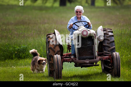 Eine ältere Frau, mit einem großen Lächeln auf ihrem Gesicht einen alten Ford Jubiläums-Traktor in Richtung der Kamera mit ihrem Hund nach hinten fahren