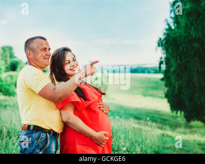 Schöne schwangere Frau im roten Kleid und ihr Mann mit den Händen auf Bauch im Freien. Mann umarmt von hinten Bauch schwanger Frau Stockfoto