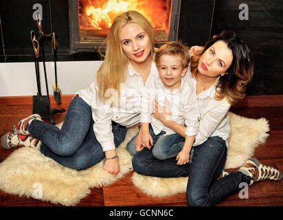 Schöne Familie bestehend aus Mutter mit Ingwer Haare, mit blonden Haaren Tochter und einen Sohn auf dem Fell Teppich am Kamin sitzen. Feuer im Kamin brennt. Stockfoto