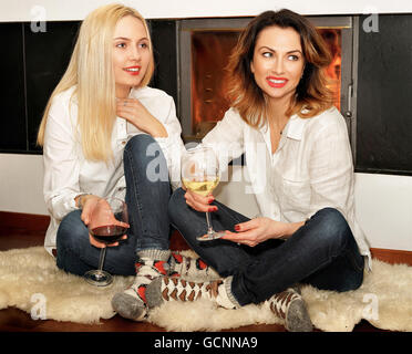 Zwei Frauen, bestehend aus einer mit Ingwer Haar und ein weiteres mit blonden Haaren auf dem Fell Teppich vor dem Kamin sitzen. Feuer im Kamin brennt. Stockfoto