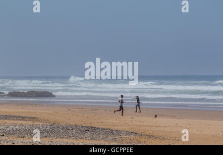 Zwei junge Männer spielen Fußball auf einem Strand mit Wellen im Hintergrund. Stockfoto