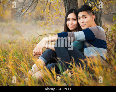 Eine junge asiatische paar genießen Sie eine romantische Zeit zusammen im Freien in einem Park im Herbst unter einem Baum sitzt und posiert für die Kamera Stockfoto