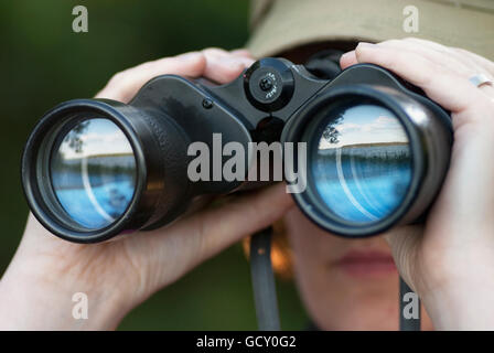 Frau, die durch ein Fernglas mit Reflexion eines Sees in den Gläsern Stockfoto