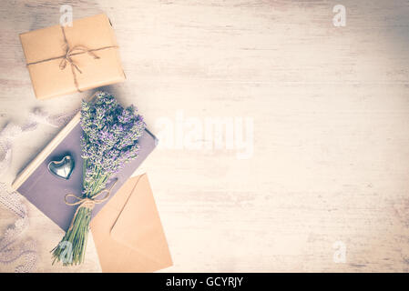 Lavendel Strauß über ein altes Buch, ein verpacktes Geschenk-Box, eine Kraft Papierumschlag und ein silbernes Herz auf einem hölzernen Hintergrund gelegt Stockfoto