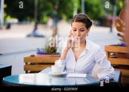 Ein gutes Geschäft zu sprechen. Fröhliche junge Geschäftsfrau telefonieren mit dem Handy und halten Tasse Kaffee im freien sitzend Stockfoto