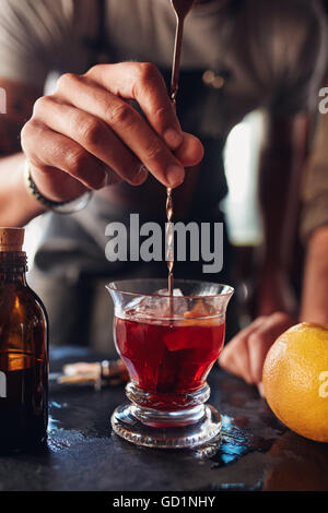 Closeup Aufnahme der Barkeeper Hand rühren Negroni Cocktail. Glas des Getränks auf Theke mit Grapefruit und Sirup Flasche. Stockfoto