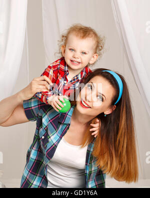 Mutter und Tochter zu Hause. Junge Mutter und Tochter umarmt und Ball spielen. Mädchen in kariertes Hemd gekleidet. Mutter tragen Stockfoto