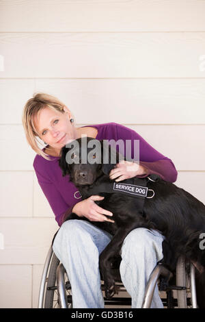 Reife Frau Rollstuhlfahrer mit ihre Arme um ihre Service-Hund, ein schwarzer Labrador, deren vorderen Pfoten, sind auf dem Schoß. Stockfoto