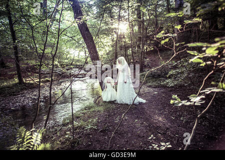 Gotischen Albtraum: eine junge blonde Frau Mädchen tragen eine altmodische weiße Hochzeit Kleid Kleid in einem Waldgebiet an einem Sommerabend, Hand in Hand mit eine gruselige Puppe gekleidet wie eine Miniatur-Braut, UK Stockfoto