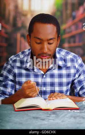Frommer Mann sitzend und Holding Rosenkranz beten und lesen aus offenen Buch auf Schreibtisch vor, Religion-Konzept Stockfoto