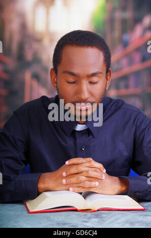 Katholischer Priester tragen traditionelle Clerical Collar Shirt sitzt mit gefalteten Händen halten Rosenkranz beim beten und lesen aus offenen Buch auf Schreibtisch vor, Religion-Konzept Stockfoto