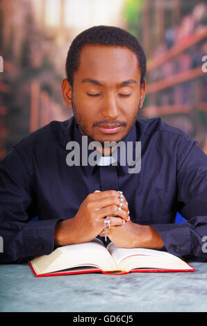 Katholischer Priester tragen traditionelle Clerical Collar Shirt sitzt mit gefalteten Händen halten Rosenkranz beim beten und lesen aus offenen Buch auf Schreibtisch vor, Religion-Konzept Stockfoto