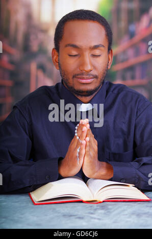 Katholischer Priester tragen traditionelle Clerical Collar Shirt sitzt mit gefalteten Händen halten Rosenkranz beim beten und lesen aus offenen Buch auf Schreibtisch vor, Religion-Konzept Stockfoto