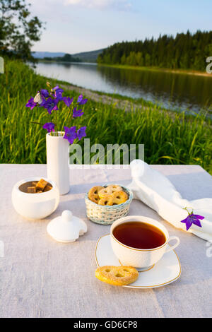 Abend Tee Sonnenuntergang Flussblick. Tasse Tee und Gebäck in kleinen Weidenkorb mit blauen Glockenblumen in Vase. Sommer-Tee-Zeit-con Stockfoto