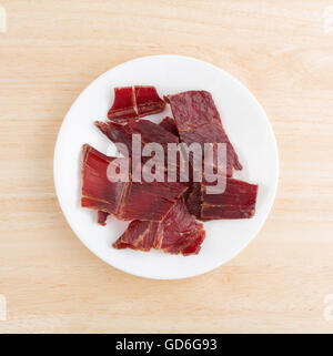 Draufsicht auf mehrere Scheiben von Laubholz geräuchert Dörrfleisch auf einem weißen Teller oben auf einer hölzernen Tischplatte. Stockfoto