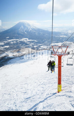 Mit dem Vulkan Yotei im Hintergrund ein weiblicher und männlicher Backcountry Skifahrer sitzen in einer Open-Air-Sessellift zum Gipfel des Mount Annupuri, im Skigebiet Niseko United auf der japanischen Insel Hokkaido.     Niseko United besteht aus vier Stockfoto