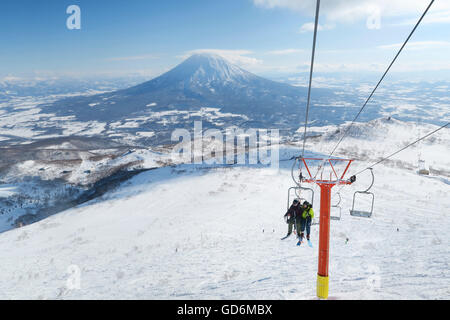 Mit dem Vulkan Yotei im Hintergrund ein weiblicher und männlicher Backcountry Skifahrer sitzen in einer Open-Air-Sessellift zum Gipfel des Mount Annupuri, im Skigebiet Niseko United auf der japanischen Insel Hokkaido.     Niseko United besteht aus vier Stockfoto