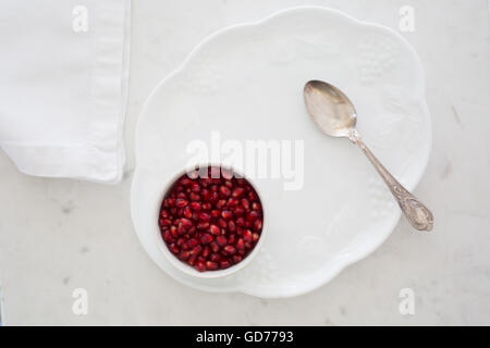 Top-Down Ansicht der Granatapfel Samen in einer kleinen Schüssel gesetzt auf einem Dessertteller auf weißem Marmor mit Serviette und silbernen Sppon. Stockfoto