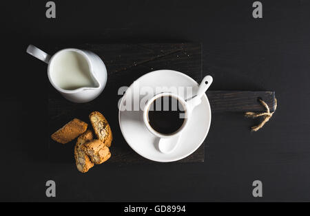 Tasse heißen Espresso, Milchkännchen mit Milch und Kekse auf dunkel rustikalen Holzbrett auf schwarzem Hintergrund, Draufsicht, horizontale compo Stockfoto
