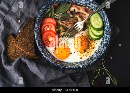 Frühstücks-Set. Pfanne mit Spiegelei mit Speck, frische Tomaten, Gurken und Brot auf schwarzem Hintergrund, Ansicht von oben Stockfoto