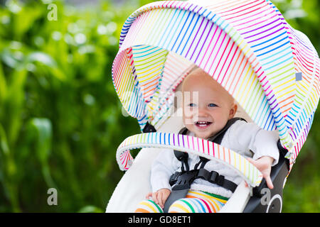 Baby Junge im weißen Pullover sitzen in weißen Kinderwagen auf einen Spaziergang in einem Park. Kind im regenbogenfarbenen Buggy. Stockfoto