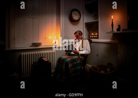 Älterer Mann sitzt auf einem Buch bei Kerzenschein mit Röhrenradio und Wolldecke auf Knien Stockfoto