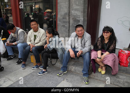 Chinesische Leute Männer und Frauen sitzen auf dem Bürgersteig vor rund um teure Geschäfte und schauen, was passiert. Qian Men Street, Beijing, China. Stockfoto