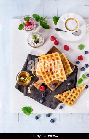 Frühstück-Thema mit belgischen Waffeln, Beeren, Honig, Kaffee und Joghurt, serviert auf Holztisch mit weißem Hintergrund. FL Stockfoto