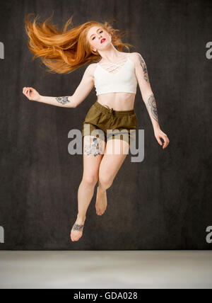 Junge Frau mit langen roten Haaren und Tätowierungen tragen grüne Hosen und springen mit Haar fließt hinter. Stockfoto