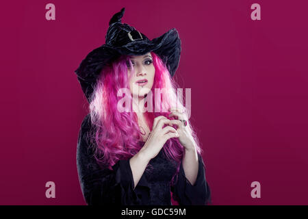 Attraktive Frau in Hexen Hut und Kostüm mit roten Haaren führt Magie auf einem rosa Hintergrund. Halloween, Horror-Thema. Stockfoto