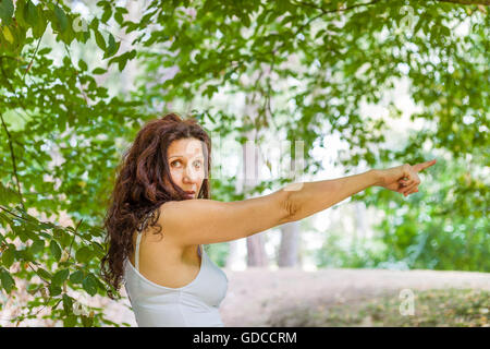 Verwirrt menopausal Frau mit Falten gekleidet in weiß und mit langen braunen welliges Haar während dem Finger der rechten Hand auf ihre linke Seite in Kamera Stockfoto
