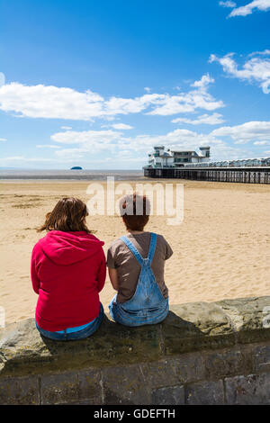 Zwei junge Frauen sassen auf der Promenade an der Wand mit Blick auf den Strand und das Grand Pier in Weston-super-Mare, Somerset, England. Stockfoto