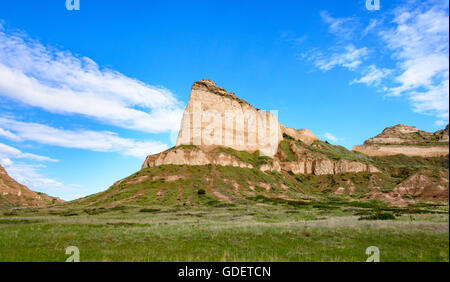 Eine hohe, massige Klippe aus geschichteten hellbraunen und weißen Steinen erhebt sich über grüne Hänge und eine große weiße Wolke bei Courthouse and Jail Rocks, Nebraska. Stockfoto