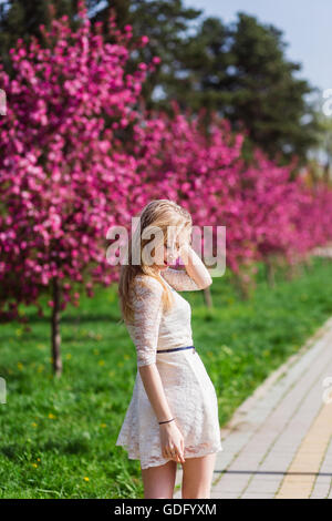 Schönes blondes Mädchen mit langen gewellten locken in einem weißen Kleid spazieren im Park zwischen den rosa Bäumen, atemberaubenden Make-up Stockfoto