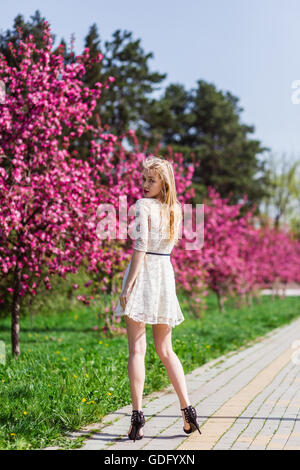 Schönes blondes Mädchen mit langen gewellten locken in einem weißen Kleid spazieren im Park zwischen den rosa Bäumen, atemberaubenden Make-up Stockfoto