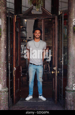 In voller Länge Portrait von stilvollen junger Mann stand an der Haustür von einem Coffee-Shop. Trendige jungen Mann am Eingang ein Café. Stockfoto