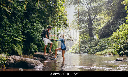 Paar im freien wandern weiter mit Mann, die Frau über Strom zu unterstützen. Junges Paar im Wald, die Überquerung des Baches. Stockfoto