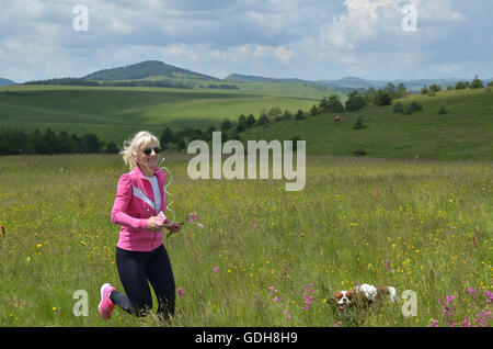 Frau läuft mit ihrem Hund mit Kopfhörern auf den Ohren über die Hügel auf Zlatibor Berg in Serbien und Stockfoto