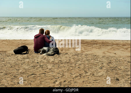 Junges Paar und ihr Hund beobachten die Wellen brechen Ont Strand Stockfoto