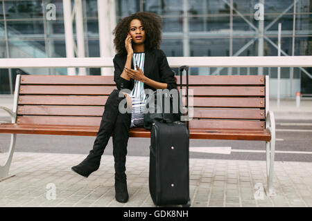 Zuversichtlich afroamerikanischen Geschäftsfrau mit Handy am Flughafen mit ihrem Koffer Stockfoto