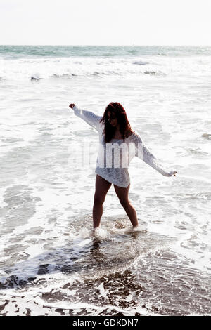 Hispanic Woman Walking In Brandung am Strand trägt weiße stricken Abdeckung über Bikini Stockfoto