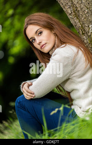 Outdoor Portrait schöne nachdenkliche trauriges Mädchen junge Frau mit roten Haaren tragen einen weißen Pullover sitzen & an einen Baum gelehnt Stockfoto